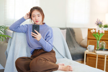 Young asian women in sweater using smartphone to chatting on social media and entertainment while sitting on sofa to relaxation and doing activity for spending time with slow life lifestyle at home