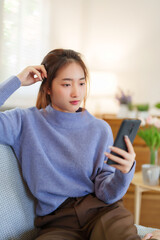 Young asian women in sweater using smartphone to chatting on social media and entertainment while sitting on sofa to relaxation and doing activity for spending time with slow life lifestyle at home