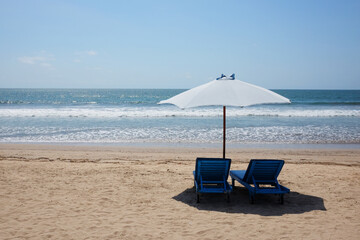 Two wooden blue seats under white umbrella outdoor on the sand and beach with blue sea in sunlight. Ocean in Bali at Indonesia