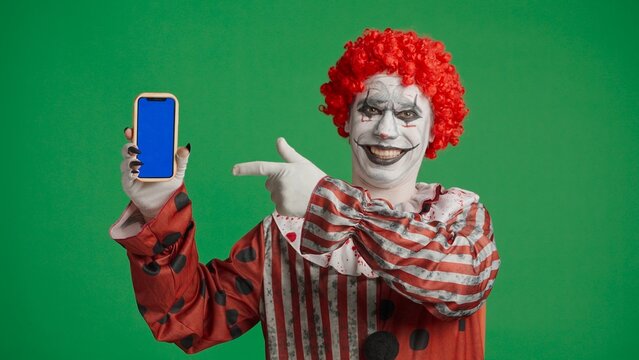 Scary halloween man with makeup dressed as clown pointing at smartphone with blue background, isolated on chroma key green screen.