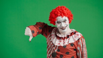 Scary halloween man clown with spooky makeup and wig dressed in costume smiles at camera shows thumb down, isolated on chroma key green screen background.