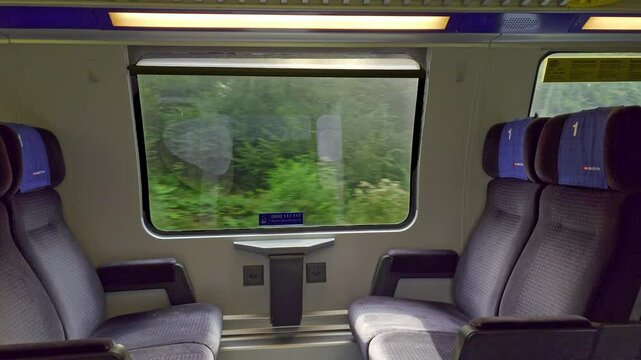 Empty first class bench seats inside moving public passenger train in Europe. Wide angle view, large windows, green summer nature landscape outside, real time, no people