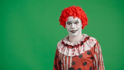 Scary halloween man clown with spooky makeup and wig dressed in costume smiles at camera, isolated on chroma key green screen background.
