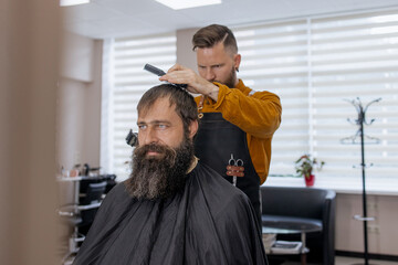 Stylish handsome barber in orange clothes and apron doing haircut to man and trimming beard in barber shop