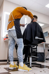 Stylish handsome barber in orange clothes and apron doing haircut to man and trimming beard in...