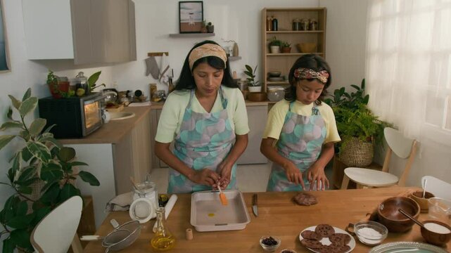 Full medium shot of Latin woman in apron and hairband pouring some olive oil into baking tray and oiling it using brush while cooking chocolate cookies together with small daughter in kitchen