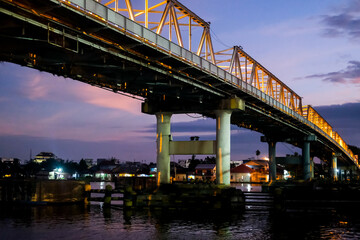 The Kapuas River Bridge in Pontianak glows beautifully at sunset, reflecting warm lights on the water. The scene captures the serene transition from day to night, showcasing urban charm.