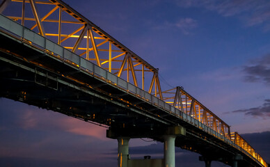 The Kapuas River Bridge in Pontianak glows beautifully at sunset, reflecting warm lights on the water. The scene captures the serene transition from day to night, showcasing urban charm.
