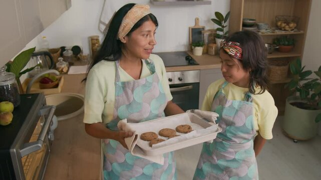 Full medium shot of smiling Hispanic mother holding hot baking tray of chocolate cookies and talking to little daughter, both wearing aprons and enjoying smell of freshly baked pastry at home