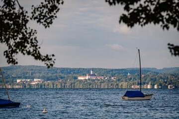 A sailboat floats on a calm lake, framed by tree branches, with a distant view of a village and a...