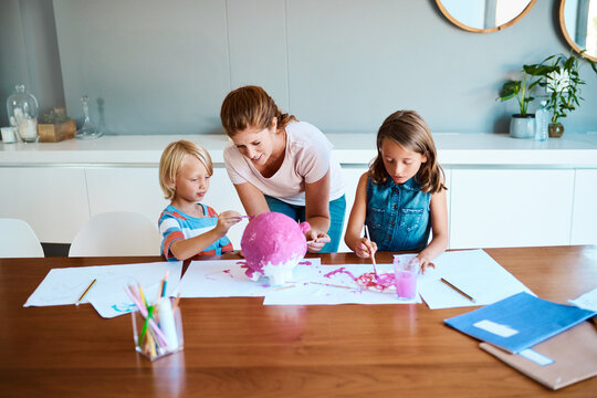 Mom, children and project in home for education, painting and design for arts and crafts. Mother, kids and creative on table for paper mache, DIY piggy bank or fun activity for development or savings