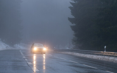 A car with headlights on is driving up the mountain road in dark and foggy winter conditions.