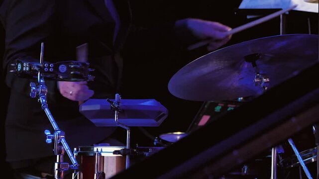 The drummer's hands pound cymbals on a dimly lit stage. A drum set with cymbals, toms and hardware. Blue and purple lights illuminate the stage. Close-up
