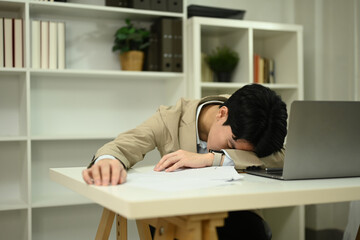 Obraz premium Tired young businessman sleeping at his office desk full of documents and laptop