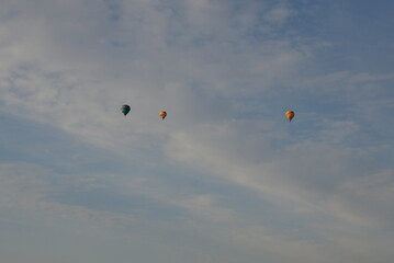 A close up on three colorful balloons flying in the cloudy sky next to some puffy clouds and a small village seen around sunrise on a Polish countryside in summer