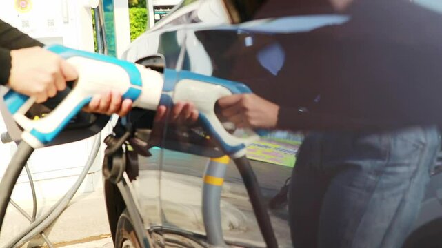 Hand young woman driving EV electric car is holding plug to her gray and black electric vehicle battery socket in a gas station : Clean energy vehicle Alternative energy is becoming popular concept.
