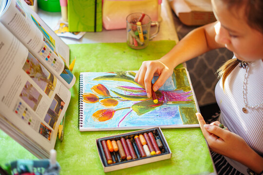 A little girl happily draws in a sketchbook at a table littered with colored pencils and pastels in a bright, sunny cheerful setting. Drawing of flowers