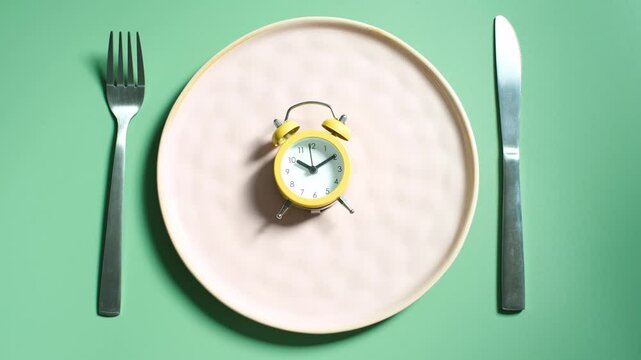 Female hand holding alarm clock against empty plate, intermittent fasting concept.