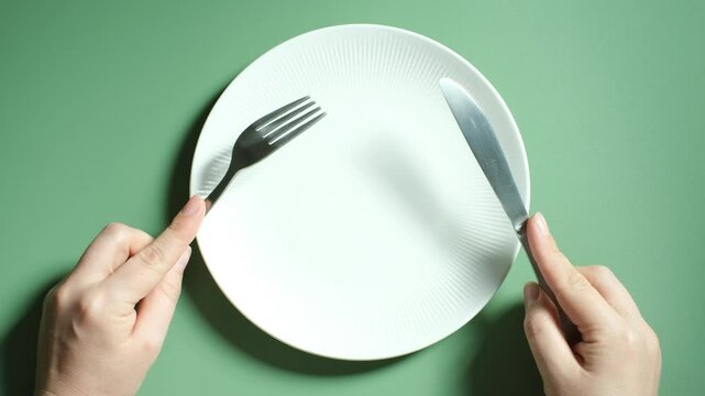 Female hands holding a fork and knife next to an empty plate on a green background close-up top view.