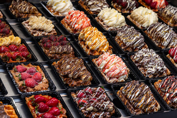 Various of traditional Belgian waffles topped with chocolate, speculoos, whipped cream and fruit display in a storefront.