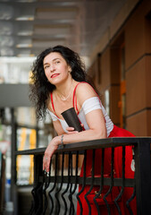 smiling brunette woman with curly hair and red lips  in red skirt  staying  with cup of coffee near vintage house in cafe