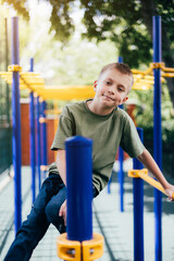 Fototapeta premium A school-age boy in a green T-shirt spends time on horizontal bars, a playground in sunny weather. Front view