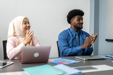 Diverse team clapping and smiling successful business presentation, muslim woman in hijab and african man in formal attire showing appreciation in a professional work setting with laptops and charts