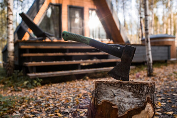An axe will be stuck into a wooden log. Harvesting firewood for the winter season. Logging on the background of a country house. Lumber, wood-cutting tools.