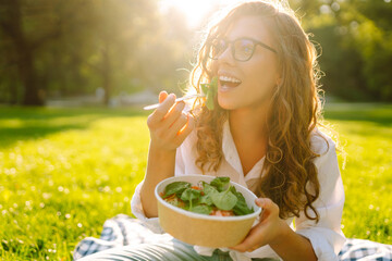 Woman enjoying a poke bowl on a sunny day in a park while sitting on a picnic blanket surrounded by...