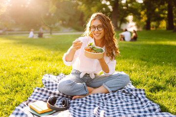 Woman enjoying a poke bowl on a sunny day in a park while sitting on a picnic blanket surrounded by...