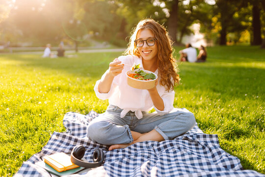 Woman enjoying a poke bowl on a sunny day in a park while sitting on a picnic blanket surrounded by green grass. Concept picnic. Vegetarian.