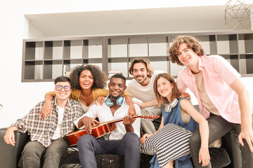 A group of multiracial teenagers playing guitar and singing a song together at an afternoon party. A group of Millennials sitting and spending time together on the holiday weekend..