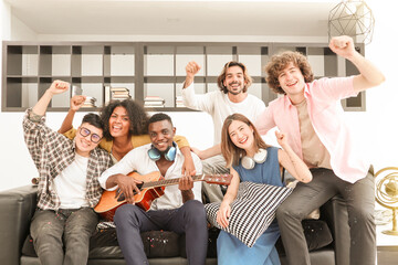 A group of multiracial teenagers playing guitar and singing a song together at the afternoon party. A group of Millennials sitting and raising their hands to celebrate the holiday weekend together.