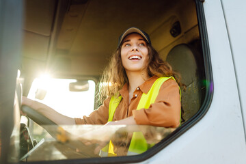 A woman with brown hair and a cap beams with joy while sitting in the driver's seat of a large...