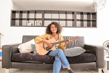 A happy, talented Black mixed-race woman is playing acoustic guitar and singing a song on a sofa in a domestic house. A beautiful female artist is practicing her singing skills before a performance.