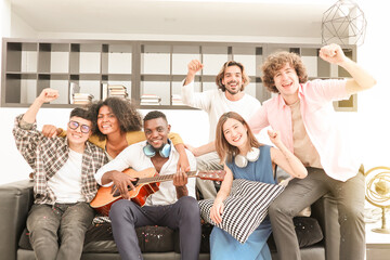 A group of multiracial teenagers playing guitar and singing a song together at the afternoon party. A group of Millennials sitting and raising their hands to celebrate the holiday weekend together.