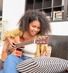 A happy, talented Black mixed-race woman is playing acoustic guitar and singing a song on a sofa in a domestic house. A beautiful female artist is practicing her singing skills before a performance.