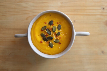 Bowl of creamy pumpkin and potato soup, with pumpkin seeds and chilli flakes toppings. Top view, wooden background.