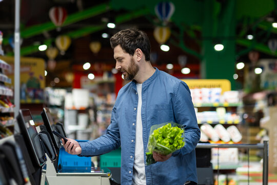 Man using self-checkout machine in busy supermarket environment holding fresh lettuce in one hand, phone in other. Image highlights modern shopping convenience, combining technology with grocery task