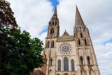 Chartres, France. The Cathedral, made of gray stone, features intricate carvings on the facade. A stunning rose window is flanked by smaller windows and statues of saints against a blue sky with