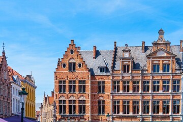 Fototapeta premium This image showcases a row of charming, old buildings with brick facades in the heart of Bruges, Belgium. The bright blue sky and white clouds highlight the colorful architectural styles.