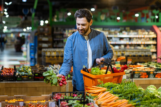 Man selecting fresh vegetables in grocery store, holding orange basket, surrounded by vibrant produce. Concept of healthy lifestyle and eco-friendly shopping in local market. Focus fresh food choice - Powered by Adobe