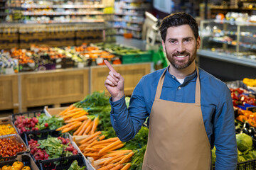 Smiling grocery store employee points at fresh vegetables in market setting. Display includes carrots, greens, and other produce. Welcoming atmosphere emphasizes healthy choices and customer service.