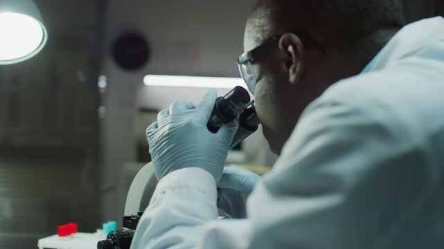 African American male serologist in gloves examining blood sample under microscope and reviewing magnified image on digital tablet while working in forensic laboratory