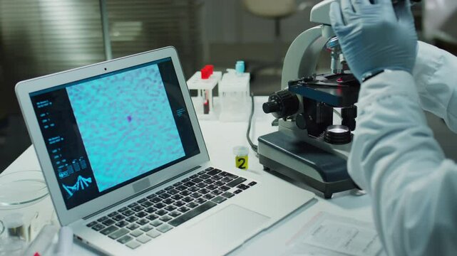 Black forensic scientist in medical gloves sitting at desk in crime lab and using digital microscope connected to laptop displaying magnified image of evidence on screen