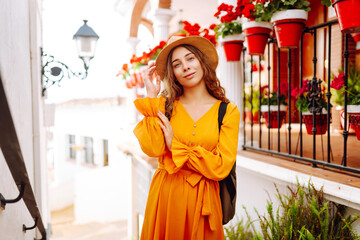 A young traveler woman in a vibrant orange dress and hat enjoys a sunny stroll through a charming...