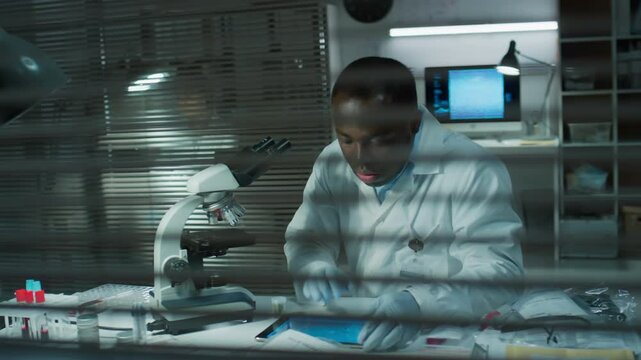 View though window blinds of African American scientist studying evidence under microscope and watching magnified image on digital tablet while working in forensic laboratory