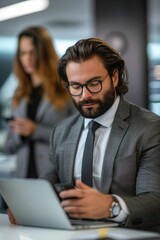 Fototapeta premium A young, bearded businessman in a suit and tie multitasks on his smartphone while sitting at his desk in an office environment.