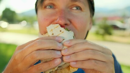 Hungry girl bites off shawarma, sitting in a street cafe against the background of a green park and road, close-up.