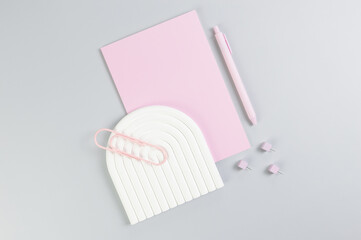 Top view of pink notebook, pink pen, paperclip, pins, white arch tray on grey background. School, office wallpaper. Flat lay, copy space.